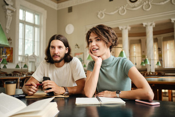 Young attractive casual students with book discussing project while studying together in library of university