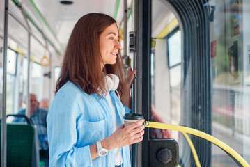 Stylish woman in blue shirt enjoying trip in the modern tram or bus, stands with cup of coffee in...