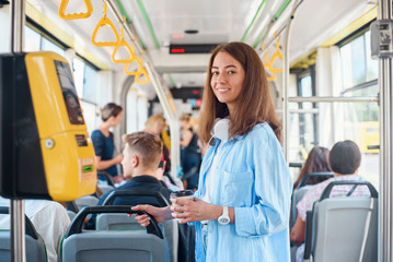 Stylish woman in blue shirt enjoying trip in the modern tram or bus, stands with cup of coffee in...
