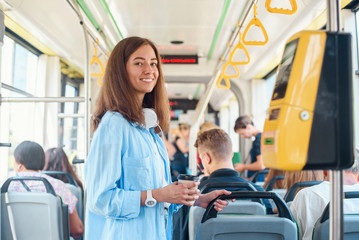 Stylish woman in blue shirt enjoying trip in the modern tram or bus, stands with cup of coffee in the public transport.