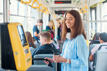 Stylish woman in blue shirt enjoying trip in the modern tram or bus, stands with cup of coffee in the public transport. - obrazy, fototapety, plakaty