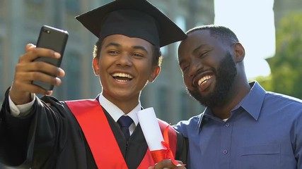 Happy emotional teenager in magisterial gown and father making selfie graduation