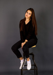 Young, beautiful girl in black clothes and white gym shoes.  A girl sits in a beautiful pose near a high chair, shooting in the studio on a plain background.
