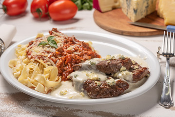Homemade pappardelle pasta with tomato sauce and beef mignon with cheese sauce in a white plate on wooden white background, soft light
