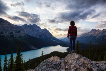 Fototapeta premium Adventurous girl on the edge of a cliff overlooking the beautiful Canadian Rockies and Peyto Lake during a vibrant summer sunset. Taken in Banff National Park, Alberta, Canada.
