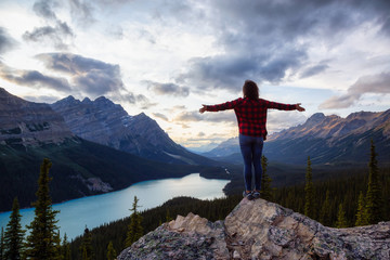 Naklejka premium Adventurous girl on the edge of a cliff overlooking the beautiful Canadian Rockies and Peyto Lake during a vibrant summer sunset. Taken in Banff National Park, Alberta, Canada.