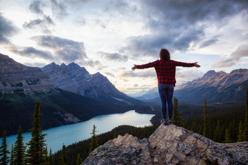 Obraz premium Adventurous girl on the edge of a cliff overlooking the beautiful Canadian Rockies and Peyto Lake during a vibrant summer sunset. Taken in Banff National Park, Alberta, Canada.