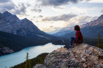 Fototapeta premium Adventurous girl on the edge of a cliff overlooking the beautiful Canadian Rockies and Peyto Lake during a vibrant summer sunset. Taken in Banff National Park, Alberta, Canada.