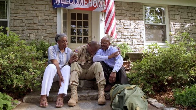 Parents hugging military son holding cell phone on front stoop