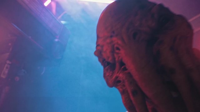 An alien with tentacles in the lower face walking on the secret laboratory in worker clothes - looking in the camera