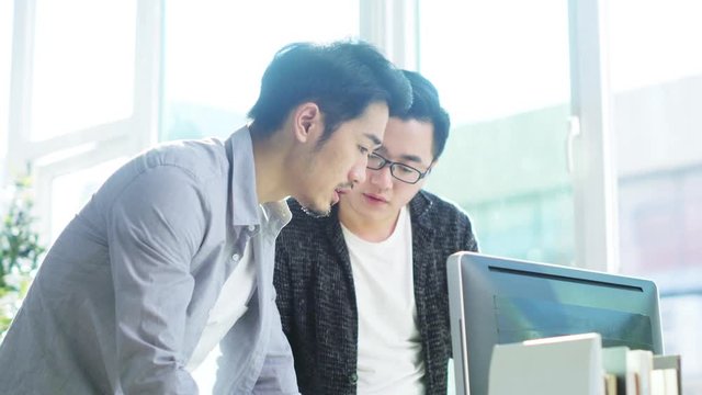 two young asian entrepreneurs standing by desk analyzing data using desktop computer