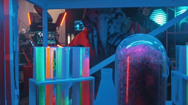 An alien with tentacles in the lower face looking around in the secret lab and touching the flasks