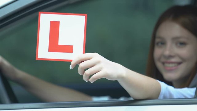 Smiling female teen showing L plate sitting car, driving license examination