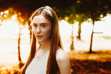 Close up of a stylish young woman looking at camera seriously against sunset. Red hair female with freckles wearing glasses outdoor .