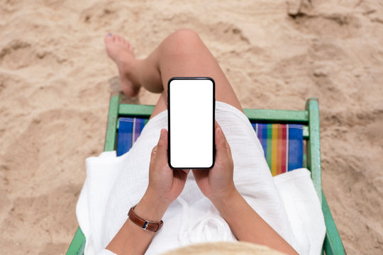 Top view mockup image of a woman holding black mobile phone with blank desktop screen while lying down on a beach chair