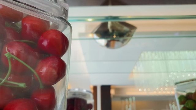 Slow Motion Slide Right To Left Shot Of A Glass Jar With Lid Filled With Decorative Plastic Cherries Sitting On An Elegant Glass Shelf With Mirror - Shot Indoors With Ceiling LED Lights