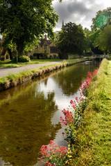 Fototapeta premium Pink flowers on the banks of the River Eye in morning sun in Lower Slaughter Cotswold District with Well landmark Gloucestershire England