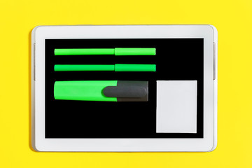 green bright felt pen, marker, pencil and paper stickers lying on the screen of the tablet pc at the yellow background. concept of office or educational stationary and gadgets