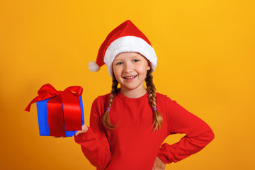 Christmas celebration. Happy little girl in santa hat holds a gift on a yellow background