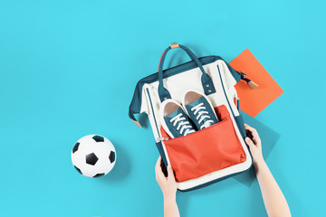 School backpack with trainers and soccer ball. Child hands holding. Flat lay on trendy light blue background with copy space. Education sports classes or activity