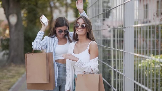 Two beautiful girls are walking on the street and holding a package from shopping centre. They are smiling, dancing and posing for a photo.