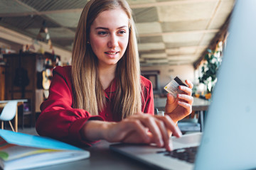 Pretty woman in red suit making online payment using plastic credit card and laptop with internet. Sitting in bright open space
