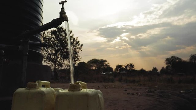 Water filling water can in refugee settlement with sunset in the background.