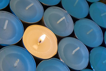 Closeup of tea lights with one being lit. A single warm orange flame between  candles in blue shadow. Christmas, mourning, condolence, memorial, funeral or cremation ceremony concept.   