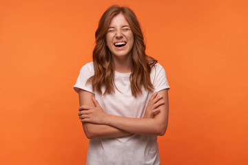 Obraz premium Portrait of cheerful young woman with red curly hair crossing arms on chest, laughing happily with closed eyes, isolated over orange background