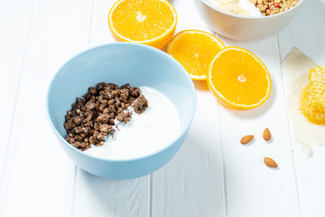 white wheat and chocolate granola with low-fat yogurt in a white bowl in a composition with a spoon, honeycombs, banana, orange on white wooden background. Healthy breakfast food. Gluten free