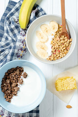 white wheat and chocolate granola with low-fat yogurt in a white bowl in a composition with a spoon, honeycombs, banana, on white wooden background. Healthy breakfast food. Gluten free