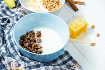 white wheat and chocolate granola with low-fat yogurt in a white bowl in a composition with a spoon, honeycombs, banana, on white wooden background. Healthy breakfast food. Gluten free