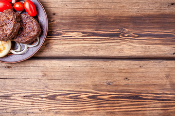 Fototapeta na wymiar Tasty grilled burger meat with vegetables - onions and tomatoes - on brown vintage style plate on wooden background. Grilled food. Copy space.