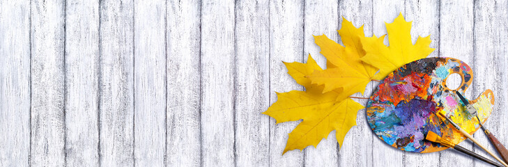 Palette for oil colors paints with brushes and palette knife on white shabby wooden table with yellow autumn maple leaves top view, art workshop concept