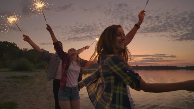 Cheerful male and female friends are running along the beach at sunset, holding sparkling fireworks and runaway lights in slow motion. Dancing and sunset party on the beach.