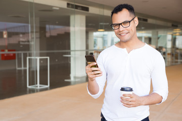 Smiling guy with smartphone looking at camera. Handsome African American man holding smartphone and paper cup of coffee. Technology concept