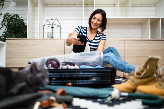 Young woman tourist packing her winter and autumn warm stuff into a big suitcase. Her beautiful Russian Blue cat lying in front. Traveling preparation concept.