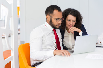 Two colleagues looking at laptop screen together. Business man and woman sitting and standing at conference table, using computer and talking. Consulting or mentorship concept