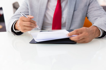 Businessman signing contract. Business man sitting at office table, holding pen, reading document. Signing agreement concept. Cropped view