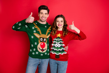 Portrait of cheerful cute two people with brunette hair embrace recommend ads show thumbs-up wear reindeer design pullover denim jeans isolated over red color background
