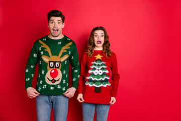 Portrait of frustrated frightened two youth people with brunette wavy hair dislike christmas tree pattern decor jumper shout omg wear denim jeans isolated over red color background