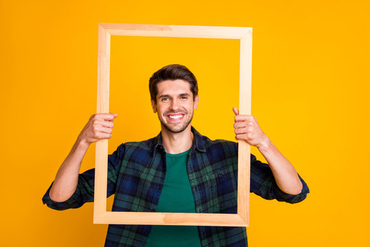 Photo of funny guy holding wooden picture frame looking through it posing wear casual checkered shirt isolated yellow color background