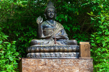 Buddha statue sitting in lotus position for worship and meditation of Buddhists in a flowering green garden with green trees and leaves. Religion and culture of the eastern countries.