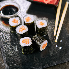 Macro shot of salmon hosomaki sushi on natural black slate plate background with selective focus. Thin maki sushi rolls with raw trout, cucumber, rice, sesame and nori closeup