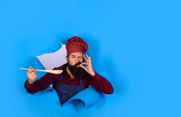 Bearded man chef preparing to cook food. Handsome chef cook with kitchen utensils. Chief man in cook uniform with wooden spoon looking through paper. Bearded cook holds wooden kitchen cooking tools