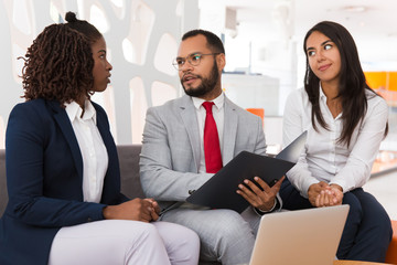 Diverse business colleagues discussing project report. Business man and woman sitting on office couch with open folder and talking. Paperwork concept