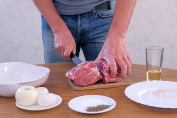 Man cutting fresh meat pork on pieces to cook shashlik, hands closeup. He is cutting on wooden board kitchen table. Prepare cook food for grilling on grill. Spices, butter and onions on table.