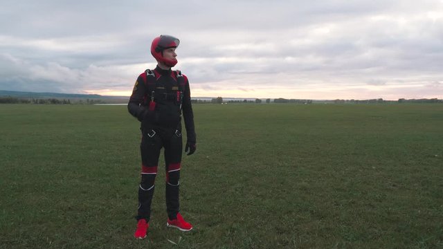 A young man in a parachutist costume stands at sunset on a green field and looks at the sky