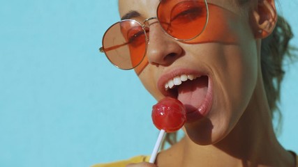 close-up of face of a young tanned laughing woman. cheerful smiling woman with red eyewear standing against blue background and sexually licking a big red lollipop. Female bright make-up for lips