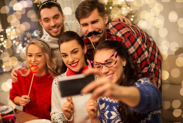 holidays and celebration concept - happy friends with party props taking selfie by smartphone at home christmas dinner